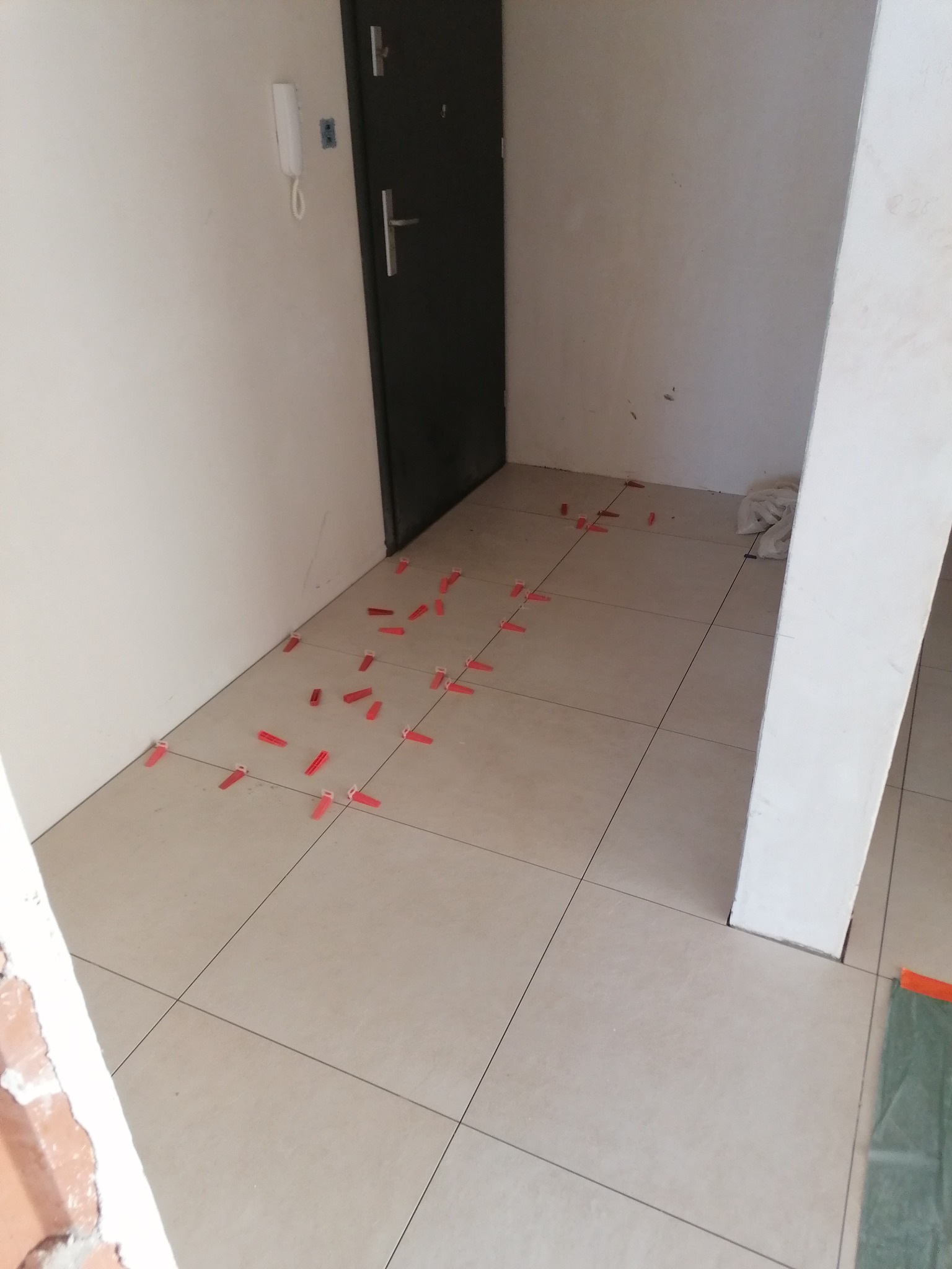 Układanie płytek podłogowych w przedpokoju, widoczne krzyżyki dystansowe i świeżo położone płytki w odcieniach beżu, przy ścianie widoczny fragment instalacji domofonowej.