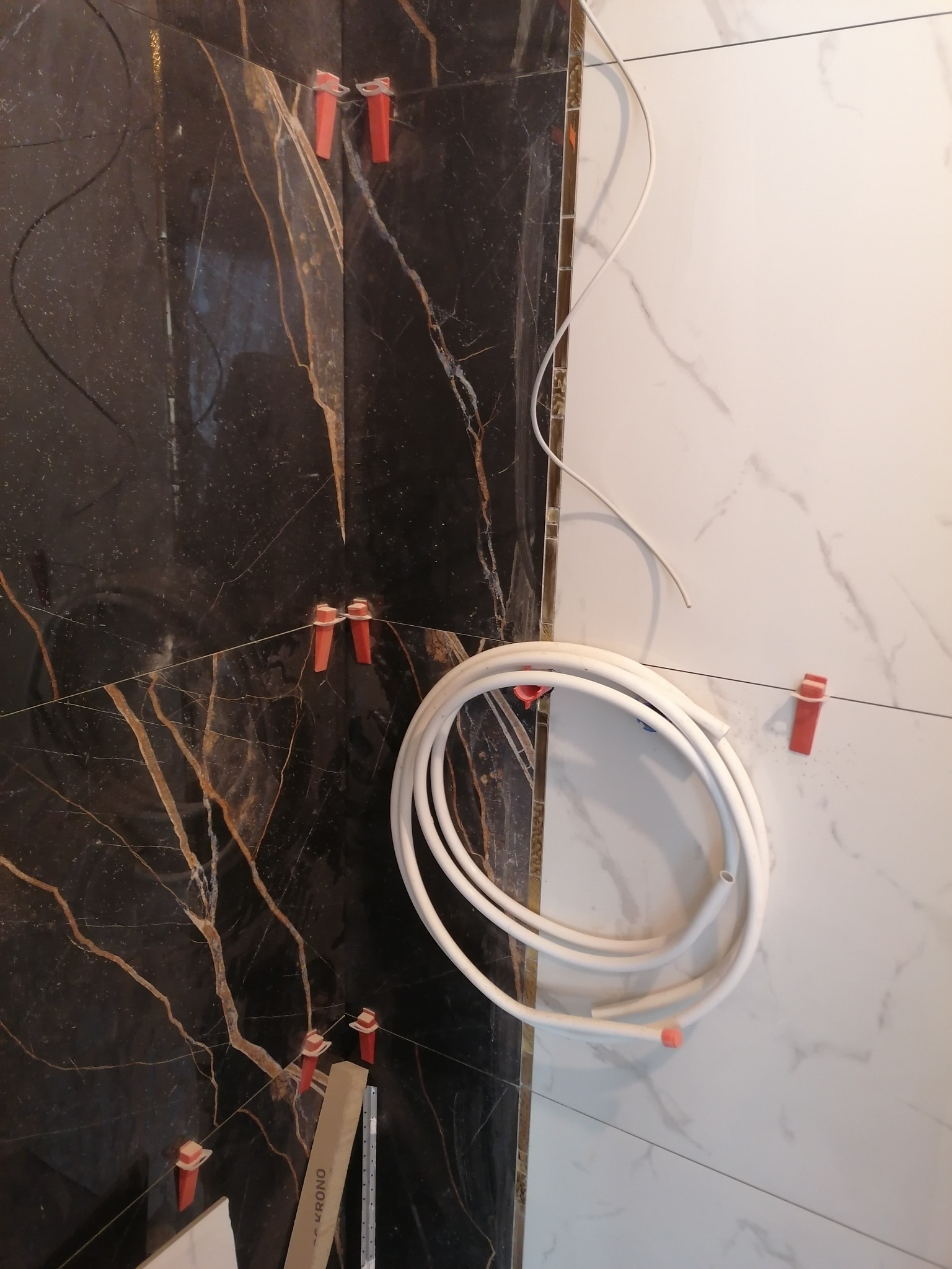 Montaż płytek imitujących marmur w łazience, widoczne płytki czarne z brązowymi żyłami i białe z szarymi żyłami, system poziomowania płytek z czerwonymi klipsami, biały przewód.