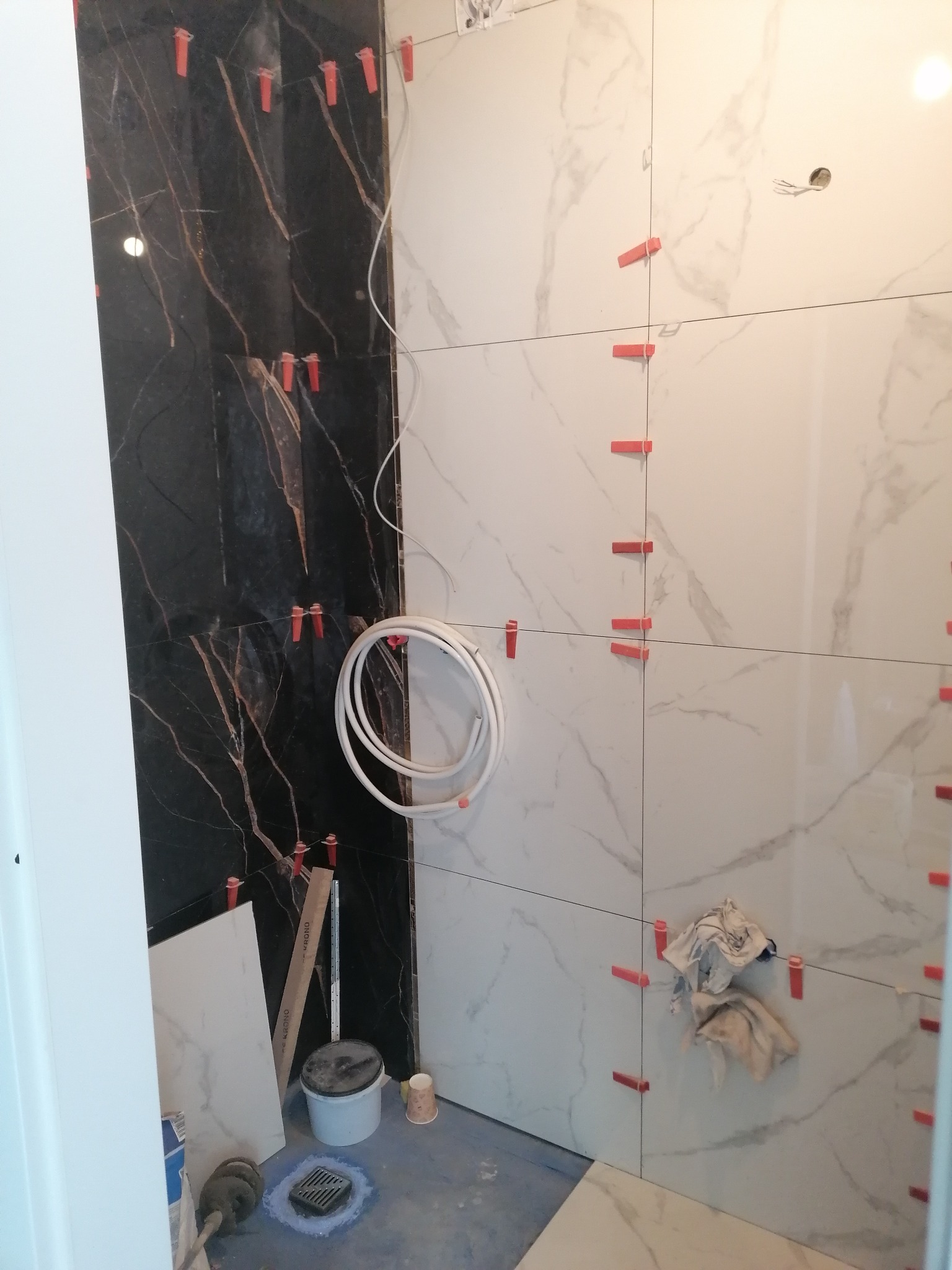 Układanie płytek imitujących marmur w łazience, widoczne czerwone krzyżyki dystansowe, czarne płytki z brązowymi żyłami, białe płytki z szarymi żyłami, oraz wiązka białych rurek.