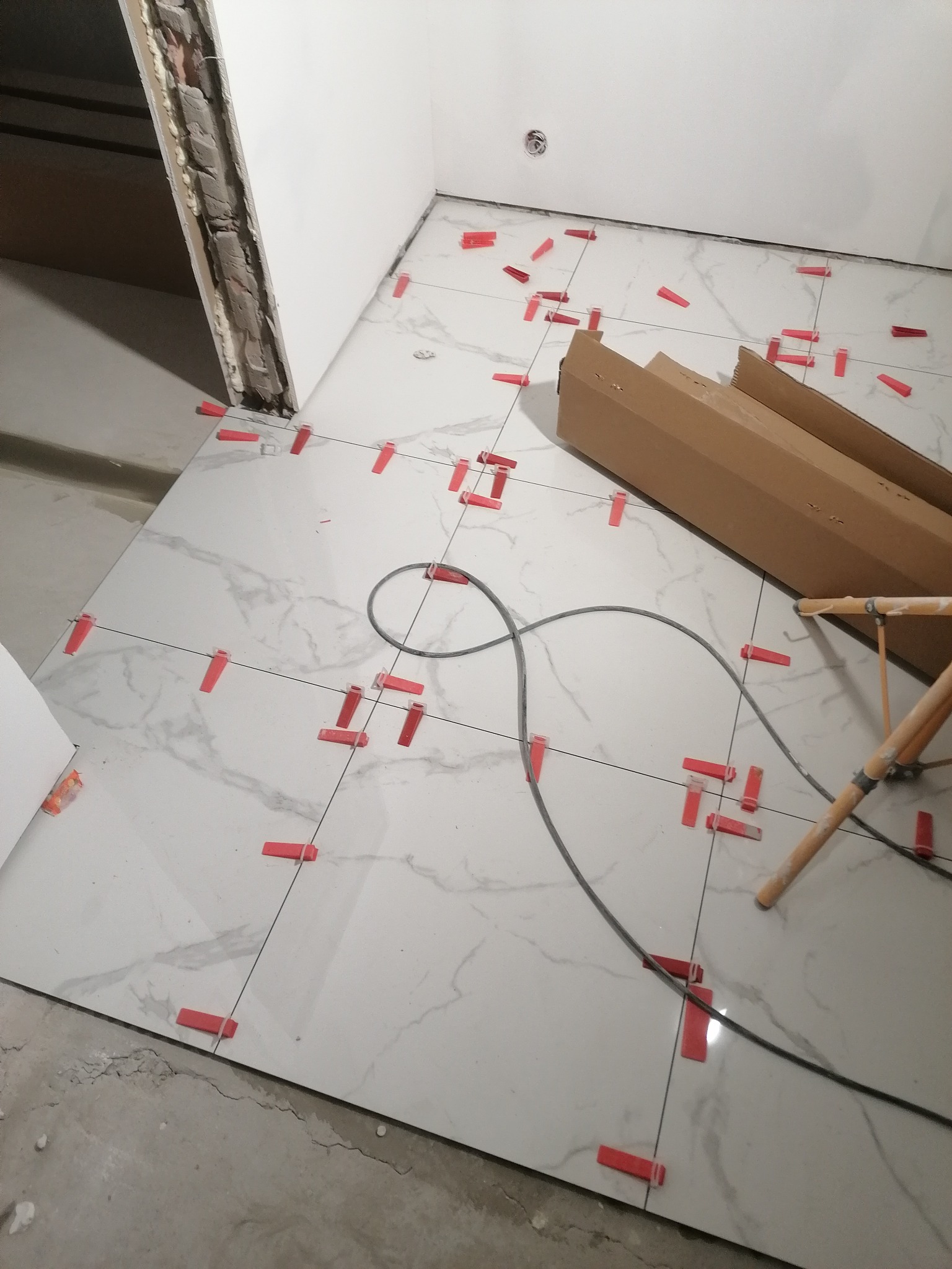 Układanie białych płytek imitujących marmur na podłodze w łazience, widoczne czerwone klipsy poziomujące, kabel i kartony po płytkach.