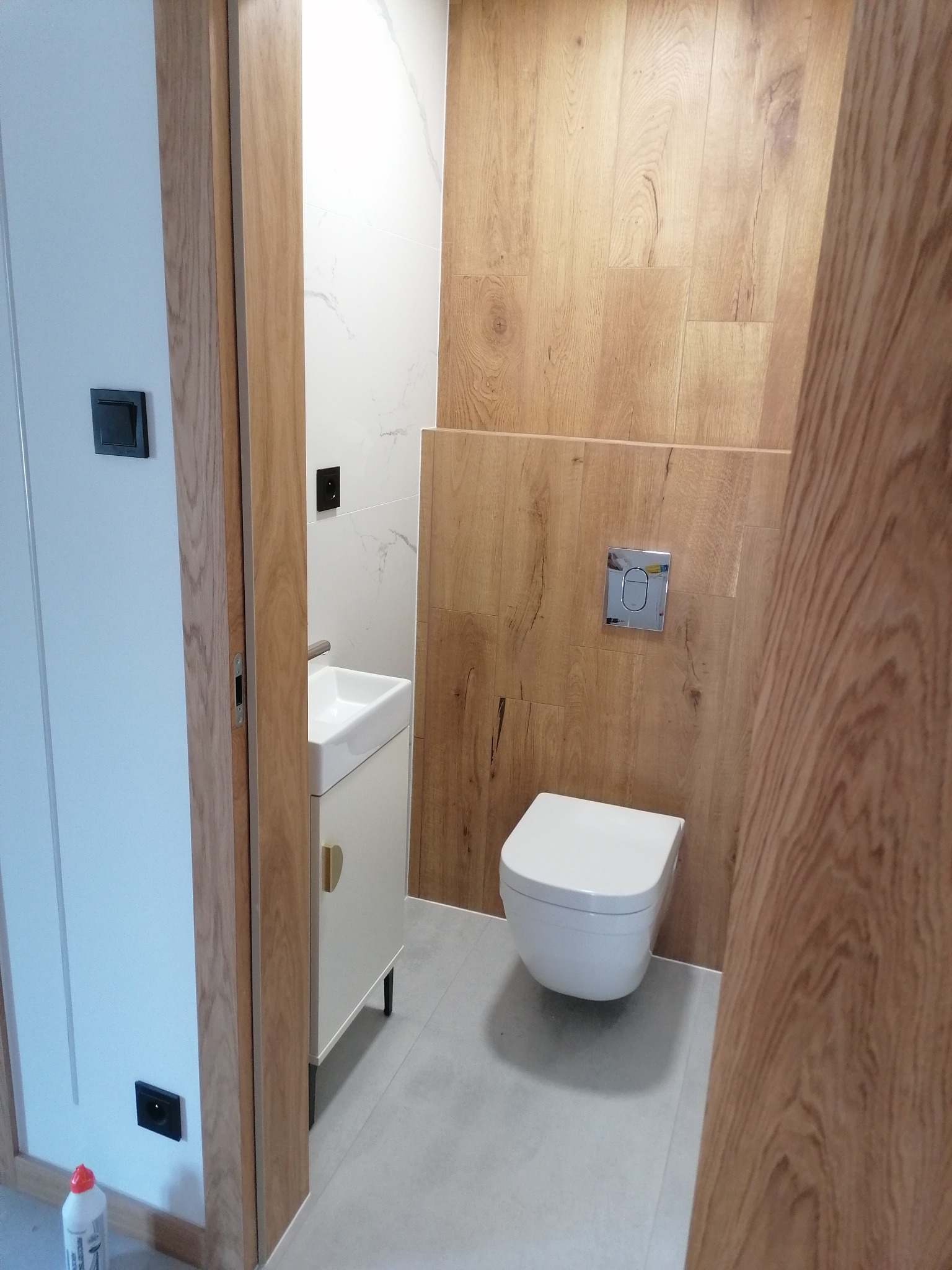 Nowoczesna łazienka z drewnopodobnymi płytkami na ścianie, wiszącą toaletą i umywalką nablatową na szafce z złotymi uchwytami, widziana zza ościeżnicy drzwi.