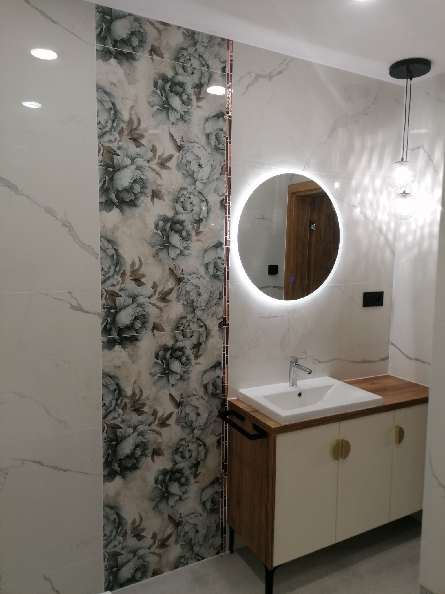 Wykończona łazienka z umywalką nablatową na szafce z drewnianym blatem i okrągłym lustrem z podświetleniem LED, płytki imitujące marmur oraz dekoracyjna ściana z motywem kwiatowym.