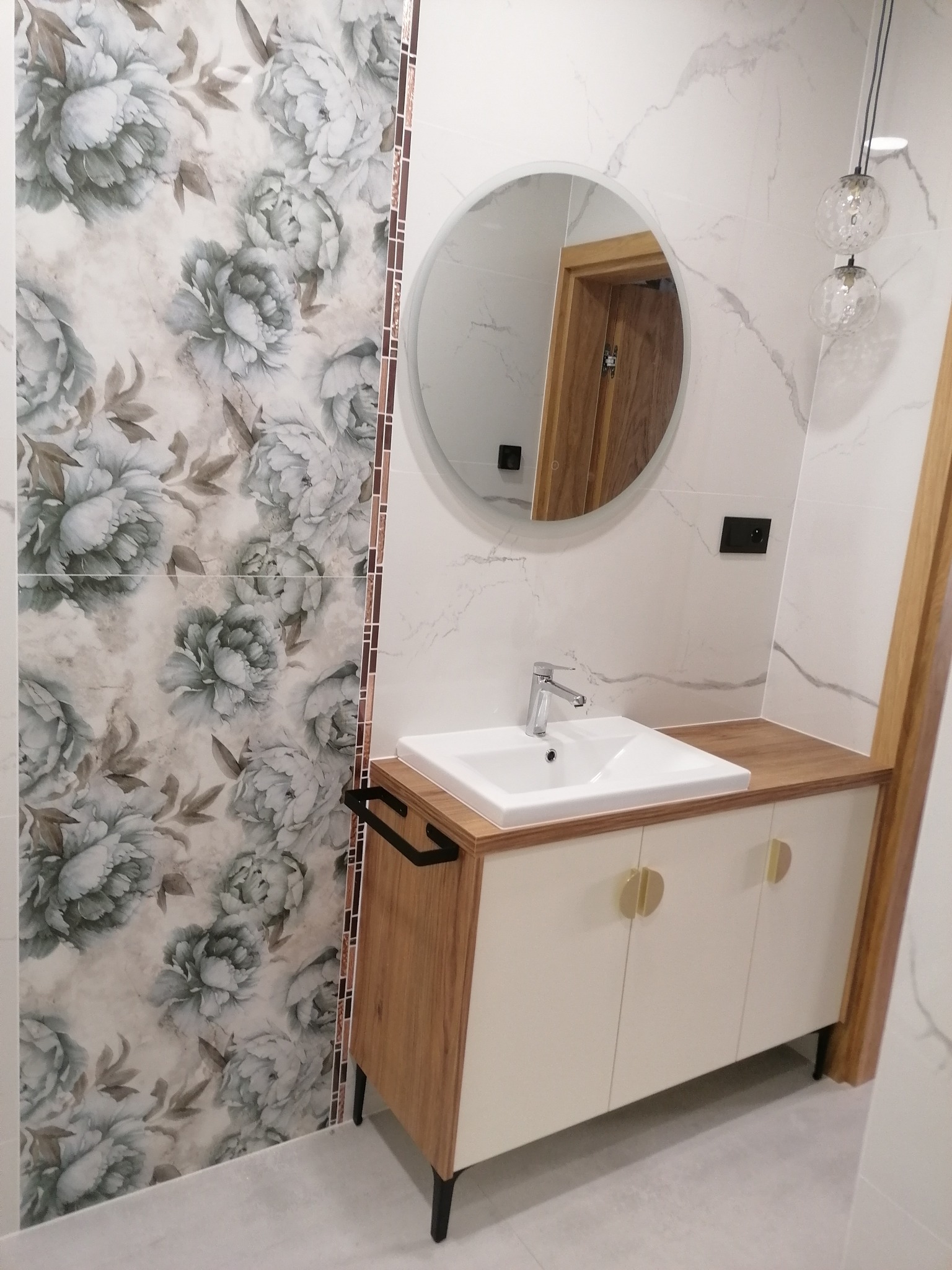 Nowoczesna łazienka z umywalką nablatową na szafce z drewnianymi akcentami i okrągłym lustrem, ozdobiona płytkami w kwiatowy wzór.