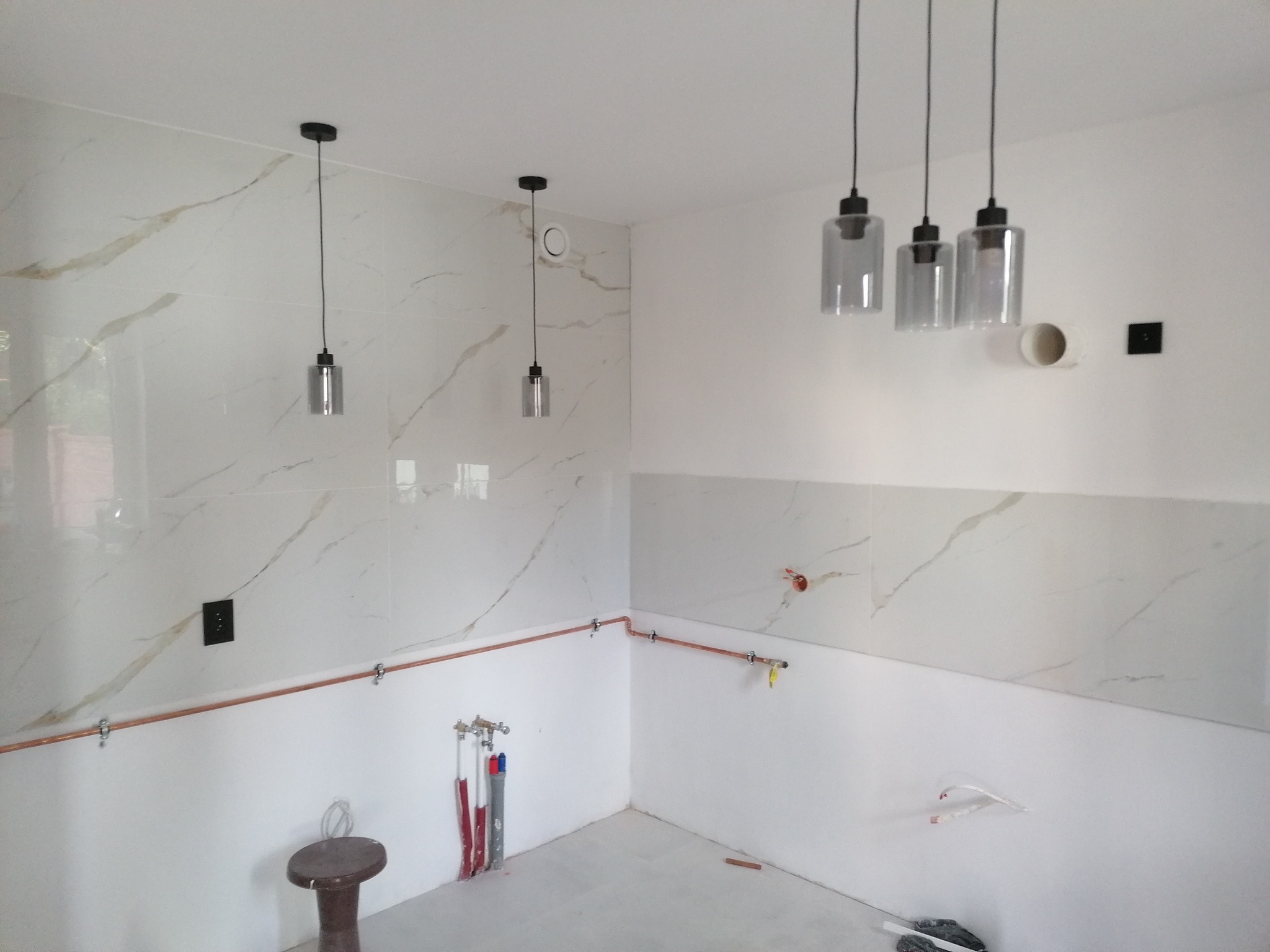 Łazienka w trakcie remontu: białe płytki imitujące marmur na ścianach, widoczne instalacje hydrauliczne i elektryczne, wiszące szklane lampy, taboret.
