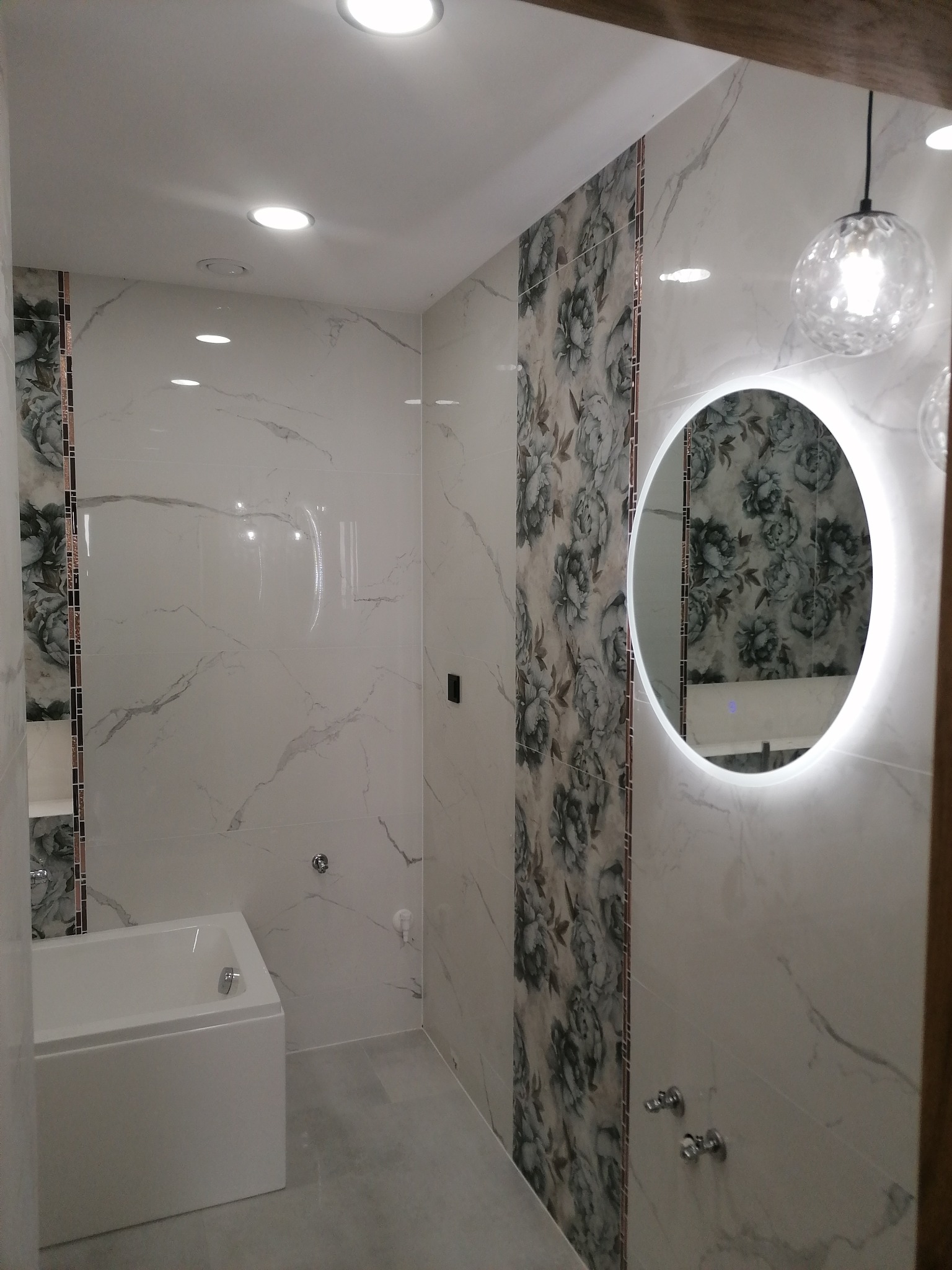 Wykończona łazienka z wanną obudowaną białymi płytkami, lustrem z podświetleniem LED i płytkami imitującymi marmur z motywem kwiatowym.