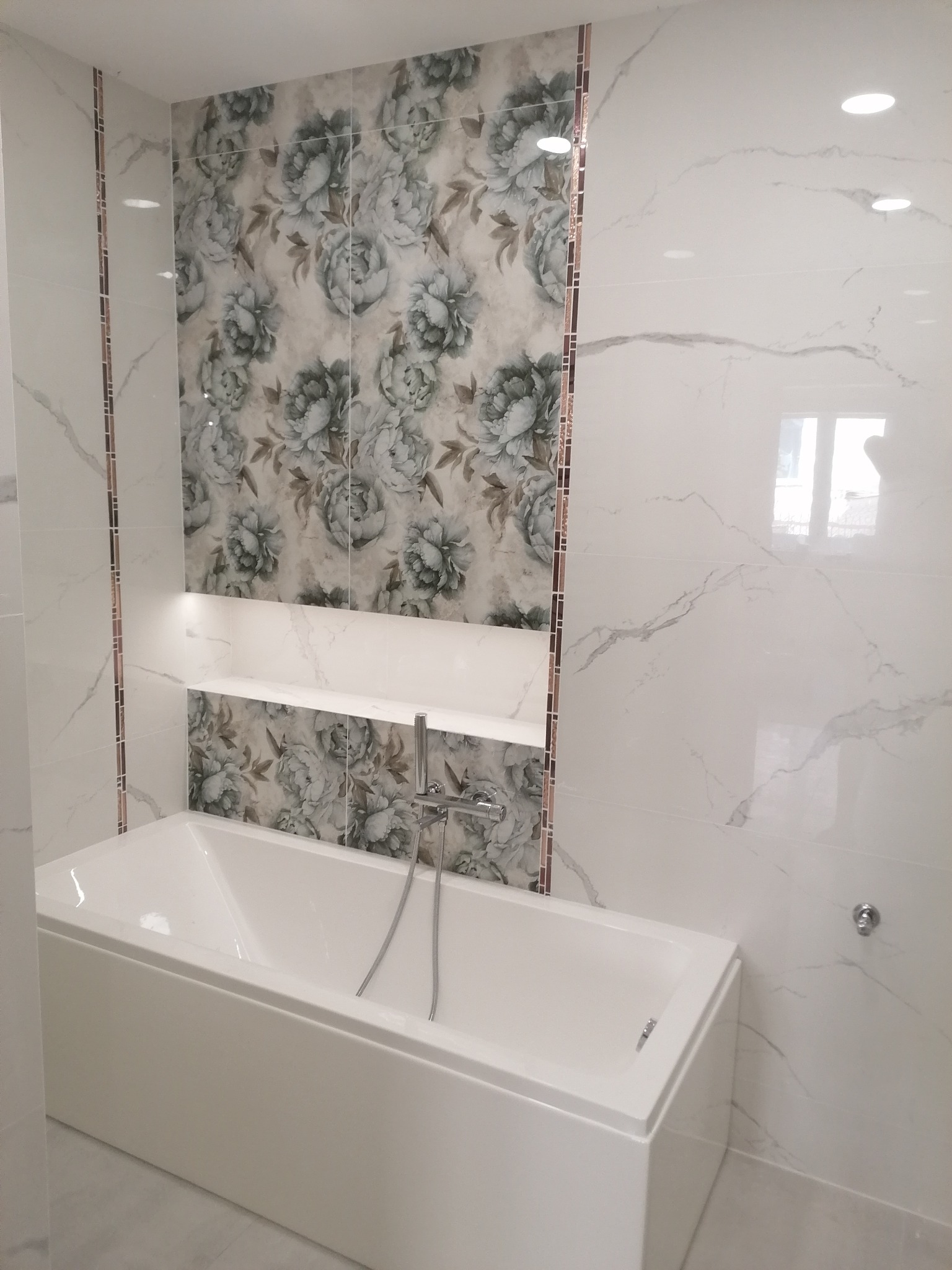Nowoczesna łazienka po remoncie: biała wanna prostokątna, ściany wyłożone płytkami imitującymi marmur i płytkami z motywem kwiatowym, półka z oświetleniem LED.