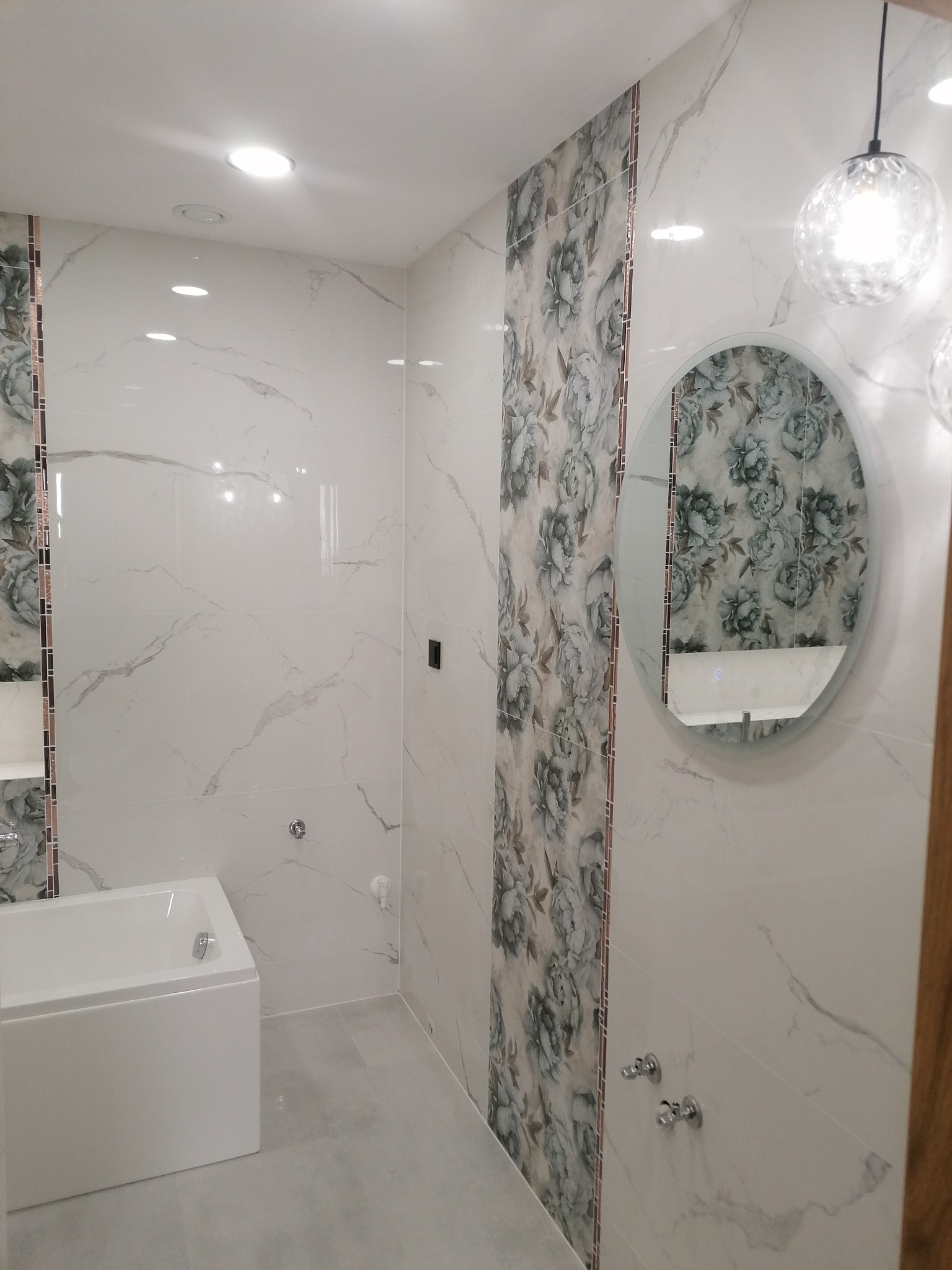 Wykończona łazienka z białą wanną, płytkami imitującymi marmur i pionowym pasem płytek z motywem kwiatowym, okrągłe lustro z podświetleniem i lampy wiszące.