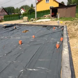 Przygotowany teren pod fundamenty z rozłożoną folią hydroizolacyjną i wystającymi rurami kanalizacyjnymi, widok na plac budowy z żółtym domem w tle.