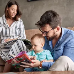 Rodzina siedząca na kanapie w salonie: kobieta z gazetą i filiżanką, mężczyzna czytający książkę dziecku, laptop na stoliku kawowym.