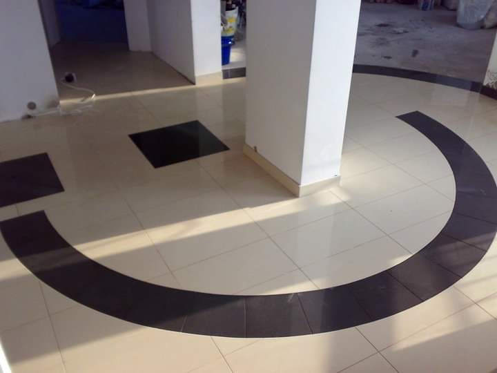 Ułożona podłoga z jasnych kwadratowych płytek, z ciemnymi wstawkami tworzącymi okrąg wokół kolumny i kwadraty w rogach, widoczne refleksy światła.