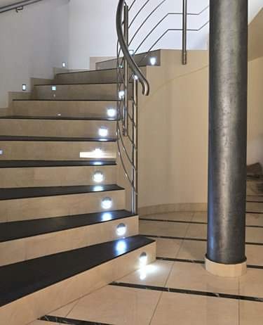 Wykończone schody z beżowych płytek z czarnymi wstawkami i punktowym oświetleniem LED w każdym stopniu, metalowa balustrada i dekoracyjny filar na jasnej, wykafelkowanej podłodze.