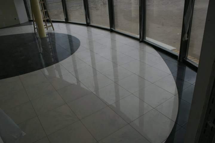 Błyszcząca podłoga wyłożona jasnymi kwadratowymi płytkami z ciemnym, półokrągłym akcentem przy oknach, odbijająca światło słoneczne.