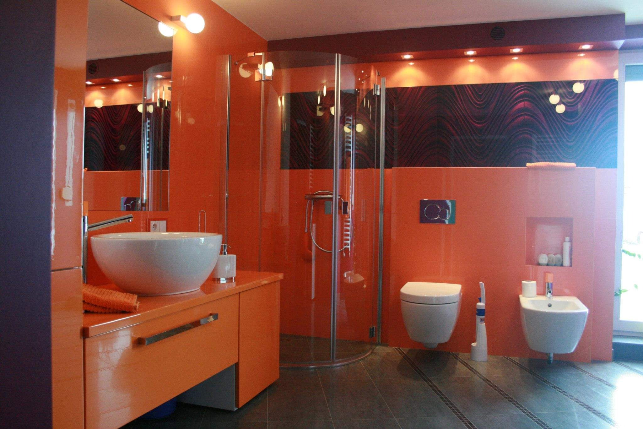 Pomarańczowa łazienka z kabiną prysznicową, umywalką nablatową, toaletą podwieszaną i bidetem; ściany wyłożone pomarańczowymi płytkami, dekoracyjny panel nad toaletą, ciemna podłoga.