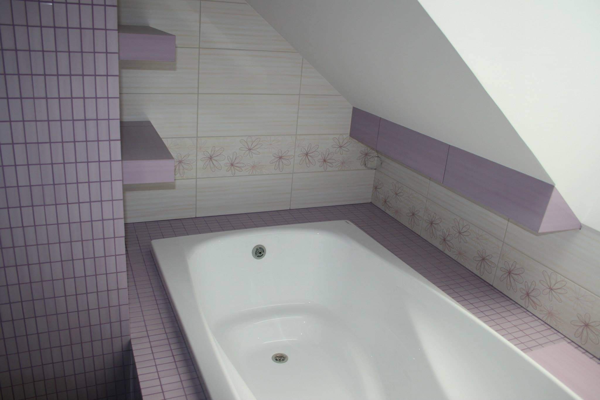 Wykończona łazienka na poddaszu z białą wanną, jasnymi płytkami z motywem kwiatowym i fioletowymi akcentami na półkach i ścianie z mozaiki.