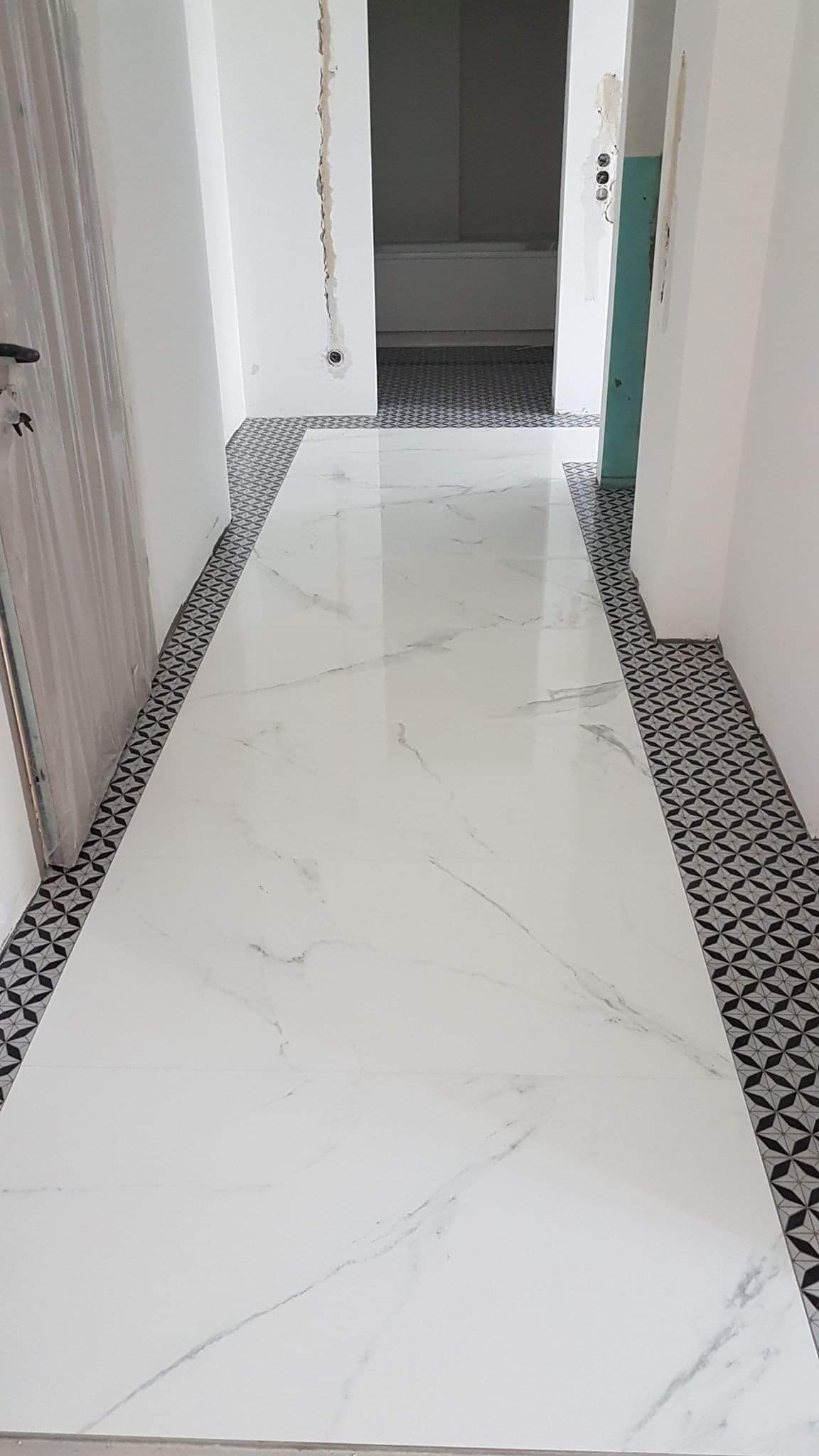 Długi korytarz wyłożony błyszczącymi, białymi płytami imitującymi marmur, obramowany dekoracyjną czarno-białą mozaiką, widoczne niedokończone wykończenia ścian.