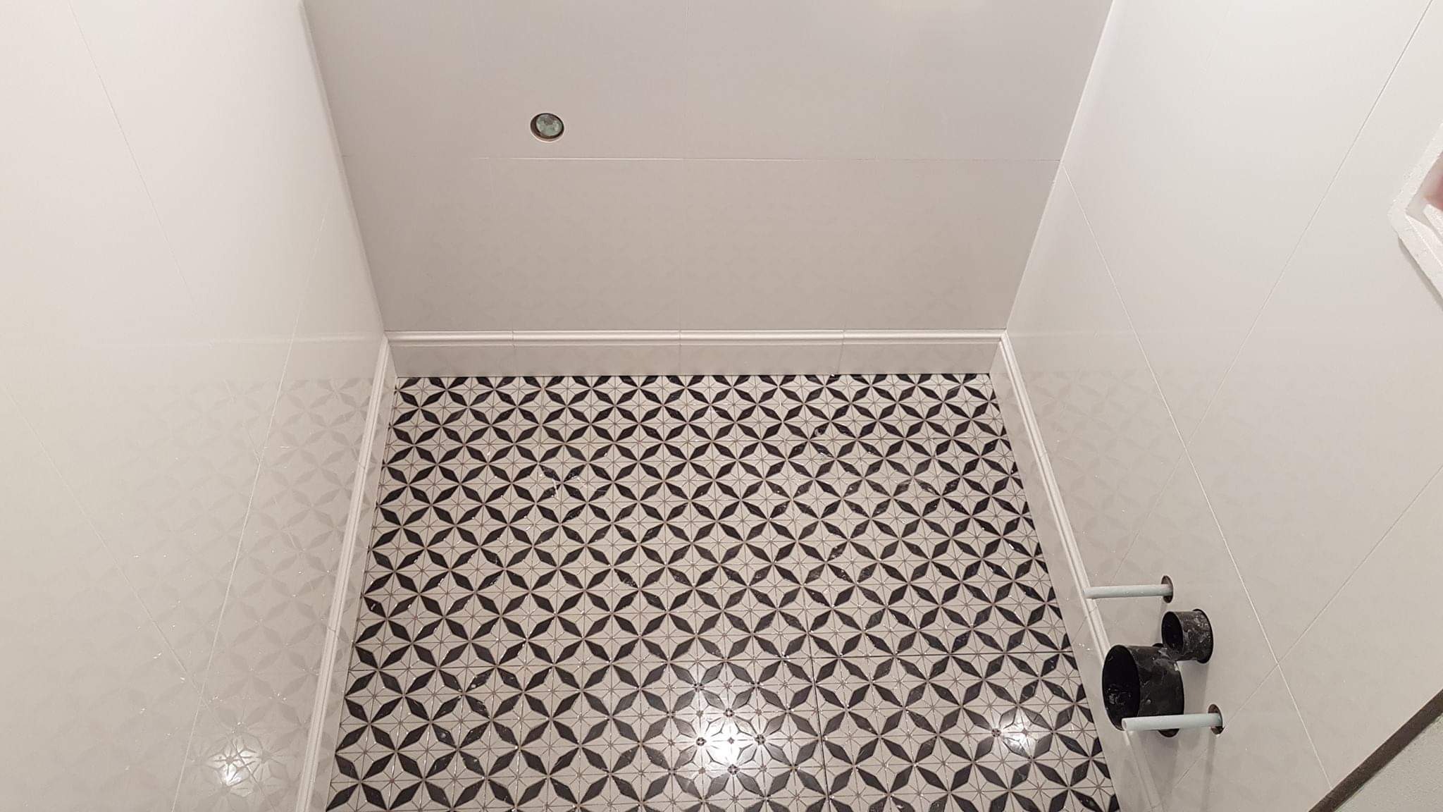 Wykończona łazienka z biało-czarną mozaiką na podłodze i białymi błyszczącymi płytkami na ścianach, widoczne rury instalacji sanitarnej.