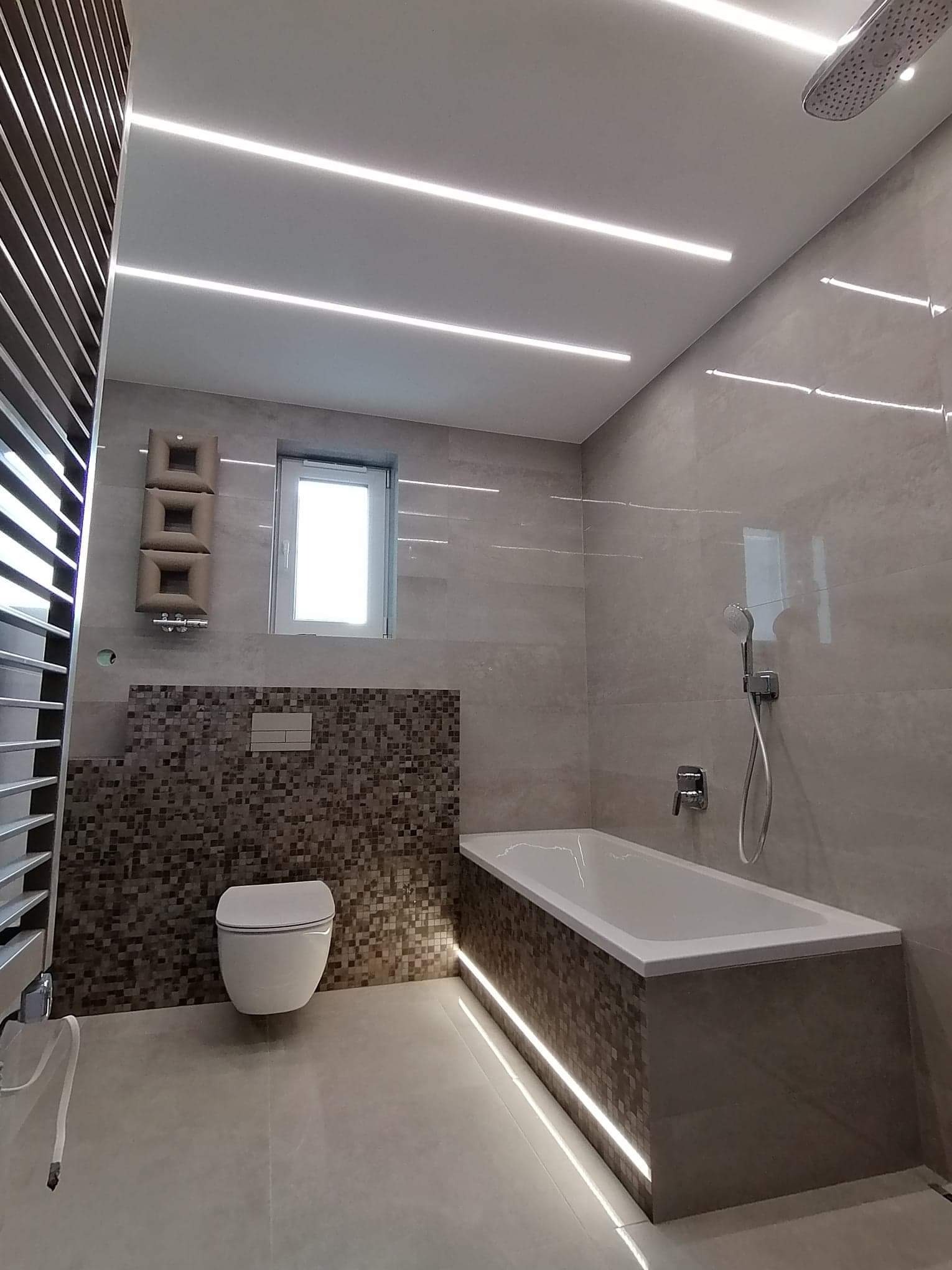 Nowoczesna łazienka z podświetlaną wanną, mozaiką na ścianie, podwieszaną toaletą i liniowym oświetleniem sufitowym.