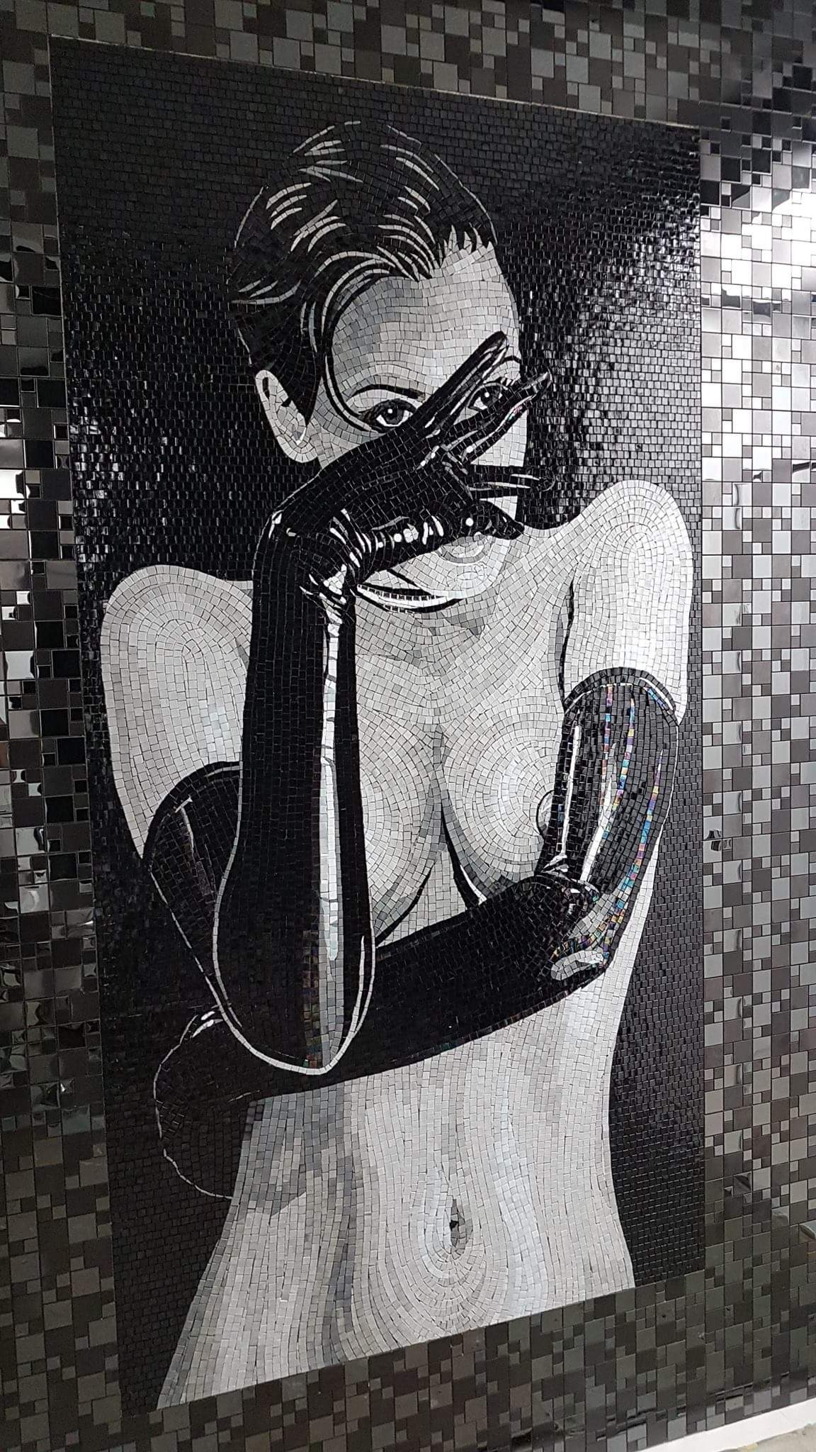 Mozaika ceramiczna przedstawiająca postać kobiety w czarno-białej tonacji, ujęcie od pasa w górę, z dłońmi w czarnych rękawicach zasłaniającymi częściowo twarz, na tle ściany wyłożonej ciemnymi...