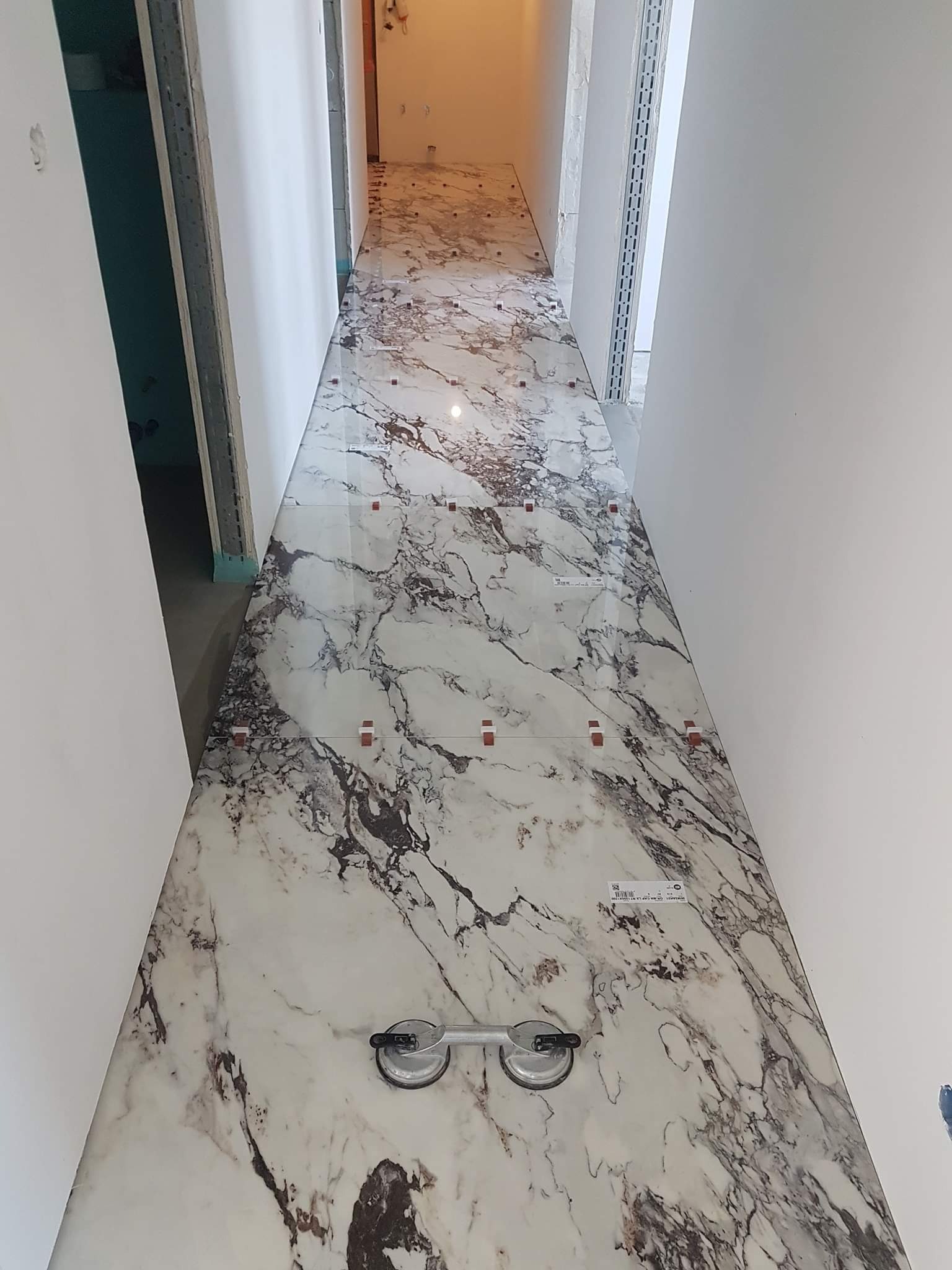 Układanie marmurowych płytek podłogowych w korytarzu, widoczne przyssawki do przenoszenia płytek i krzyżyki dystansowe.