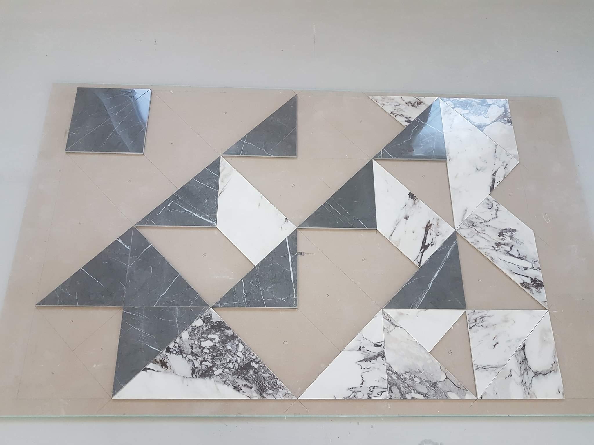 Układanka z trójkątnych i kwadratowych płytek imitujących marmur w odcieniach szarości i bieli, ułożona na beżowej powierzchni z zaznaczonym rysunkiem pomocniczym podziału.