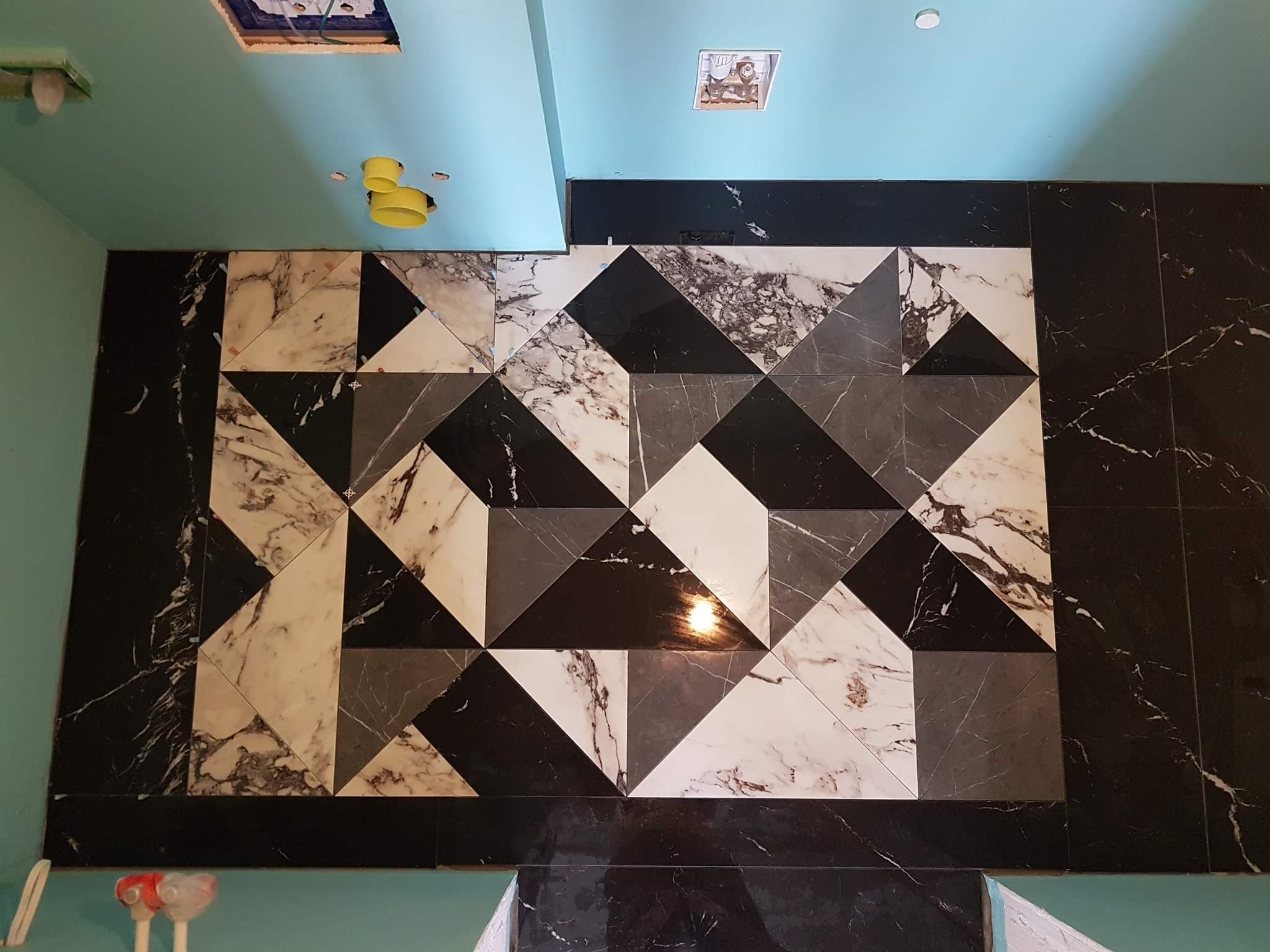 Mozaika z trójkątnych płytek marmurowych w odcieniach bieli, szarości i czerni na ścianie łazienki z niebieskim sufitem i widocznymi instalacjami elektrycznymi.