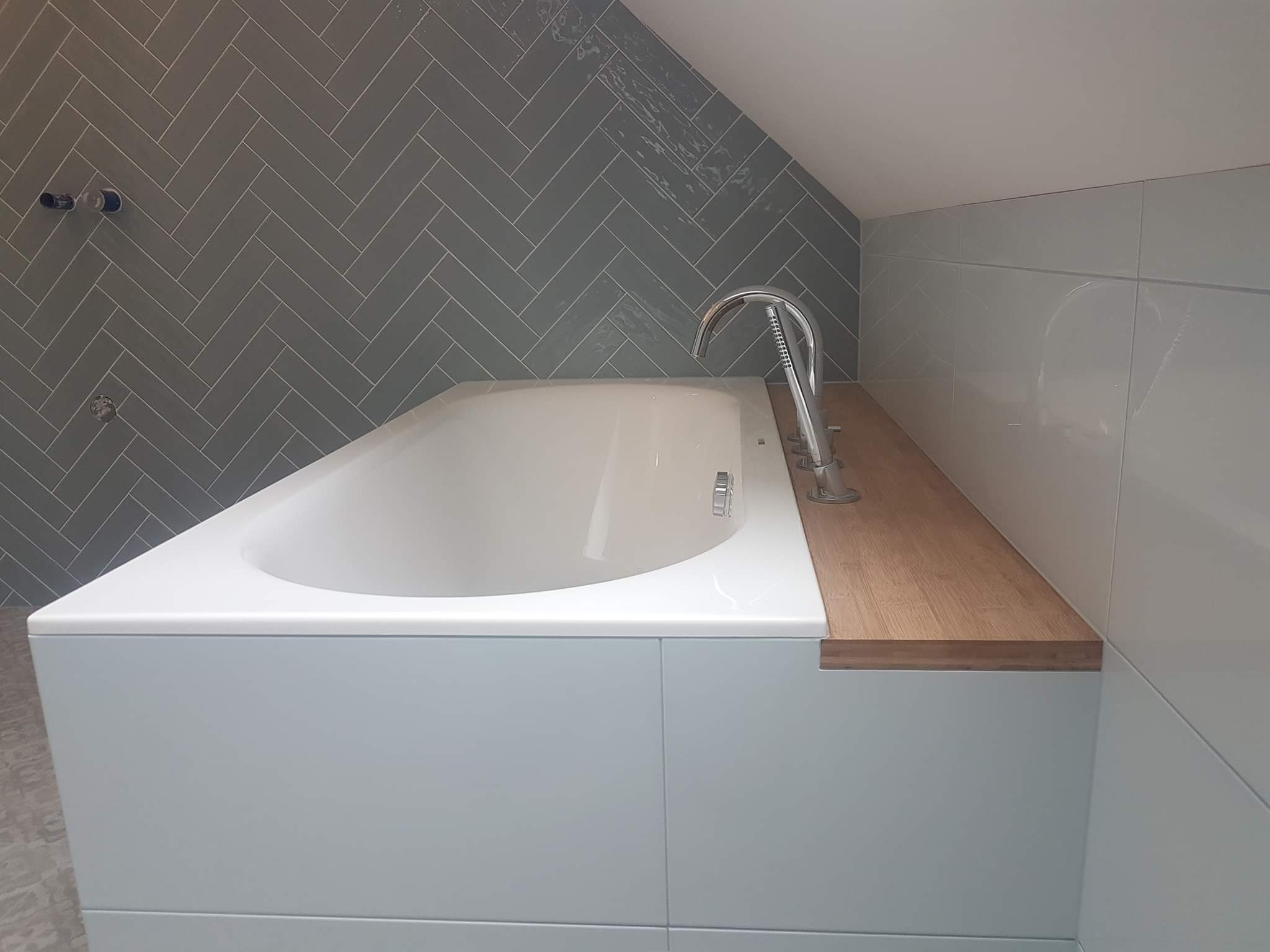 Wykończona łazienka z wanną obudowaną jasnoniebieskimi płytkami, drewnianym blatem i szarą ścianą wyłożoną płytkami w jodełkę. Widoczne chromowane bateria wannowa oraz fragment poddasza.