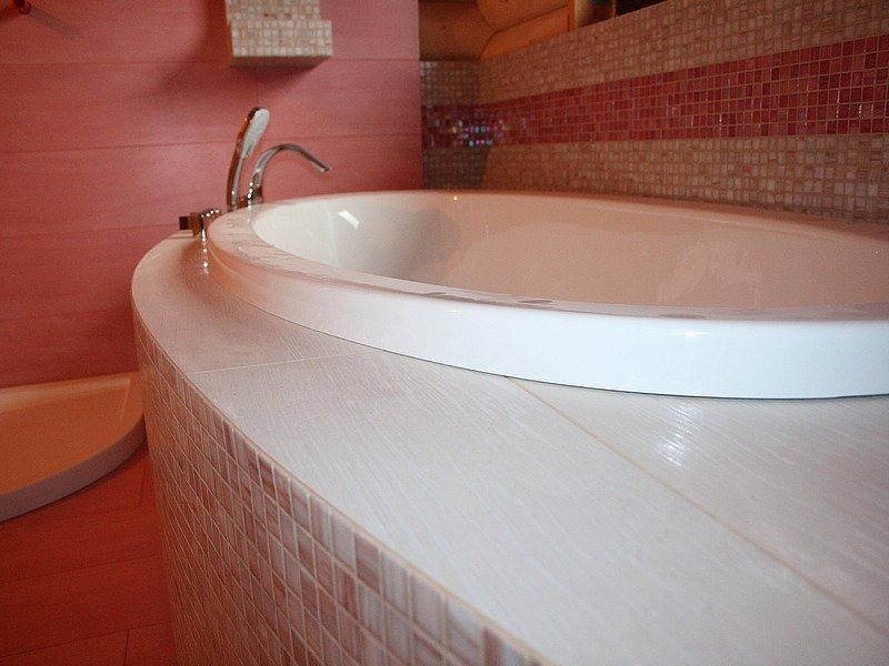 Zaokrąglona obudowa wanny wyłożona mozaiką w odcieniach różu i beżu, z blatem imitującym drewno, w tle bateria i ściana w kolorze pudrowego różu.