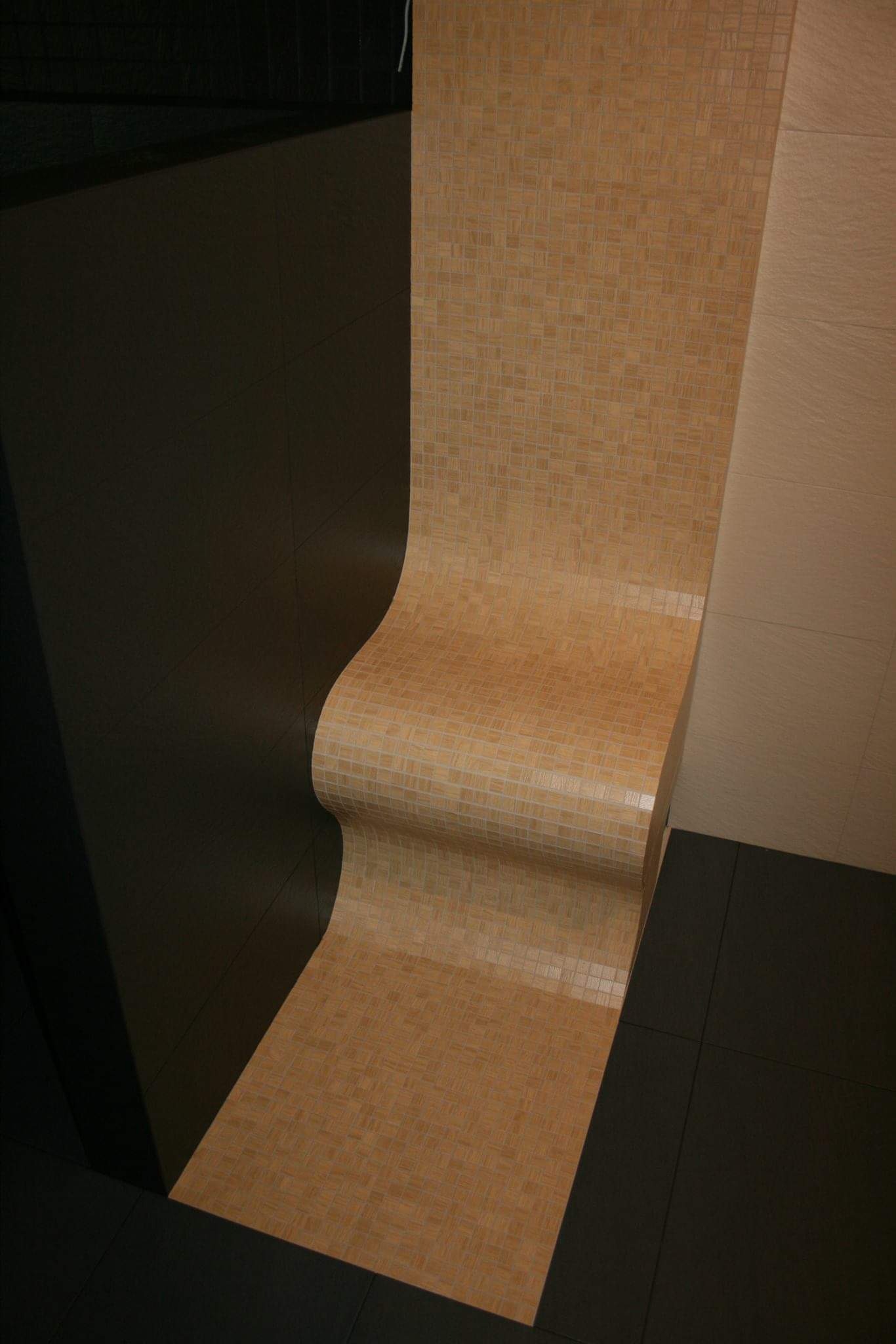 Nietypowe wykończenie ściany mozaiką ceramiczną w kolorze beżowym, z zaokrąglonymi krawędziami, przylegające do ciemnej, gładkiej ściany i jasnych płytek ceramicznych na ścianie obok oraz ciemnej...