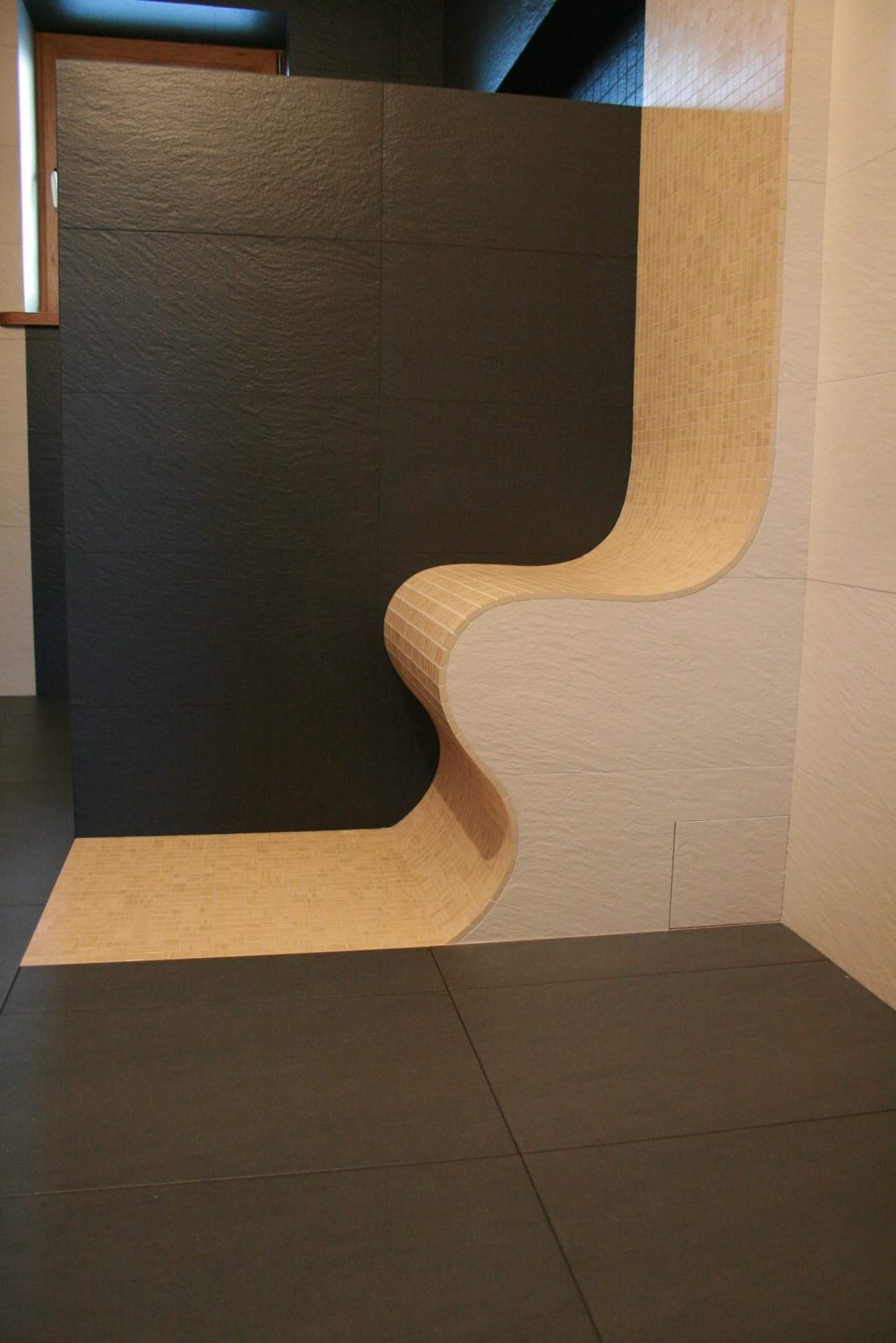Nowoczesna łazienka z czarnymi płytkami na ścianie i podłodze, oraz oryginalnym, falistym elementem z mozaiki w odcieniach beżu i bieli.