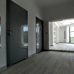 Korytarz z trzema parami drzwi w odcieniach szarości, w tym jedne szklane przesuwne, z widoczną metalową klapą rewizyjną na ścianie i drewnopodobną podłogą.