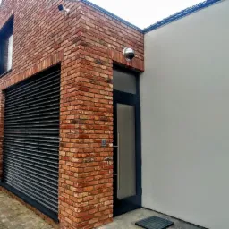 Nowoczesny dom z cegły klinkierowej z czarnymi żaluzjami zewnętrznymi i ciemnymi drzwiami wejściowymi, kamera monitoringu nad wejściem, widok z zewnątrz.