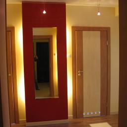 Tomasz Kowalczyk - Nowoczesny przedpokój z podświetlanym lustrem w czerwonej ramie i drewnianymi drzwiami z jasnym panelem pośrodku.