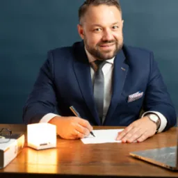 Uśmiechnięty doradca w garniturze, piszący na kartce przy biurku z laptopem i lampką. Profesjonalny wizerunek doradcy finansowego.