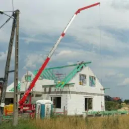 Budowa domu jednorodzinnego w trakcie, z użyciem dźwigu z wysięgnikiem podającym materiały na dach, widoczna konstrukcja dachu z zielonych łat, ściany z białego bloczka, przenośna toaleta na placu...