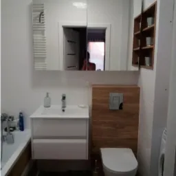 Nowoczesna łazienka z białymi płytkami, szafką z umywalką, toaletą z drewnianą obudową spłuczki i lustrem z grzejnikiem na ręczniki, odbicie osoby w lustrze.