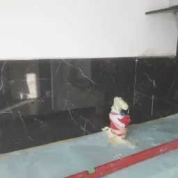 Częściowo wykończona ściana z czarnych płytek imitujących marmur w kuchni, zabezpieczona folią i poziomnicą, widoczne krzyżyki dystansowe i wiadro owinięte taśmą malarską.