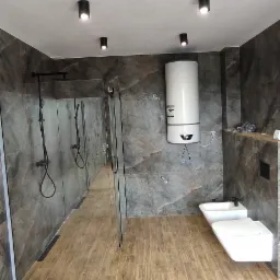 Nowoczesna łazienka z kabiną prysznicową typu walk-in, marmurowymi płytkami ściennymi, drewnopodobną podłogą, białym bojlerem i podwieszaną toaletą z bidetem, oświetlona punktowymi lampami sufitowymi.