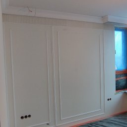 Ściana z dekoracyjnymi panelami i listwami w trakcie wykańczania. Widoczne otwory na gniazdka, zabezpieczona podłoga i drabina malarska.