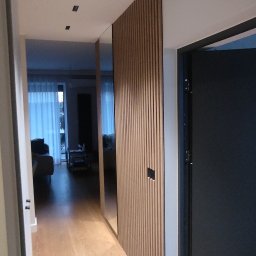 Nowoczesny przedpokój z drewnianą ścianką działową i lustrem. Jasne panele podłogowe, minimalistyczne oświetlenie sufitowe. Widok na salon przez otwarte drzwi.