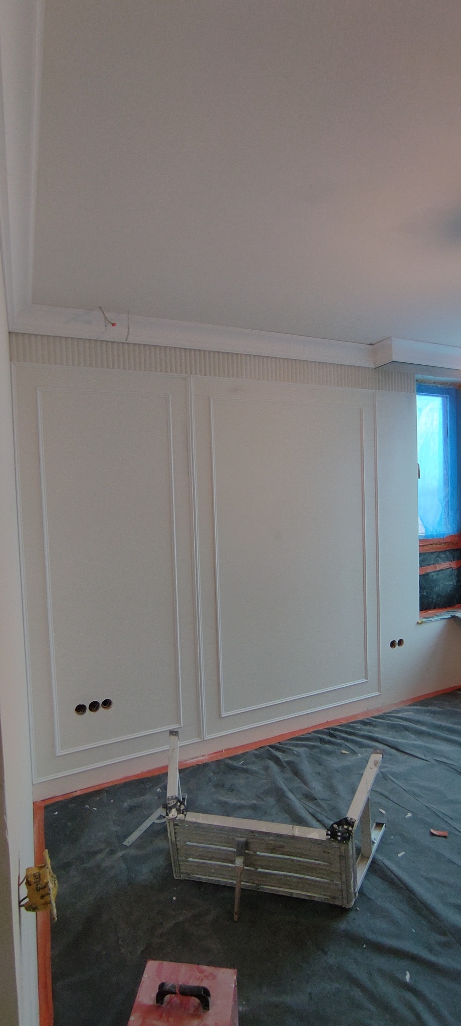 Ściana z dekoracyjnymi panelami i listwami w trakcie wykańczania. Widoczne otwory na gniazdka, zabezpieczona podłoga i drabina malarska.