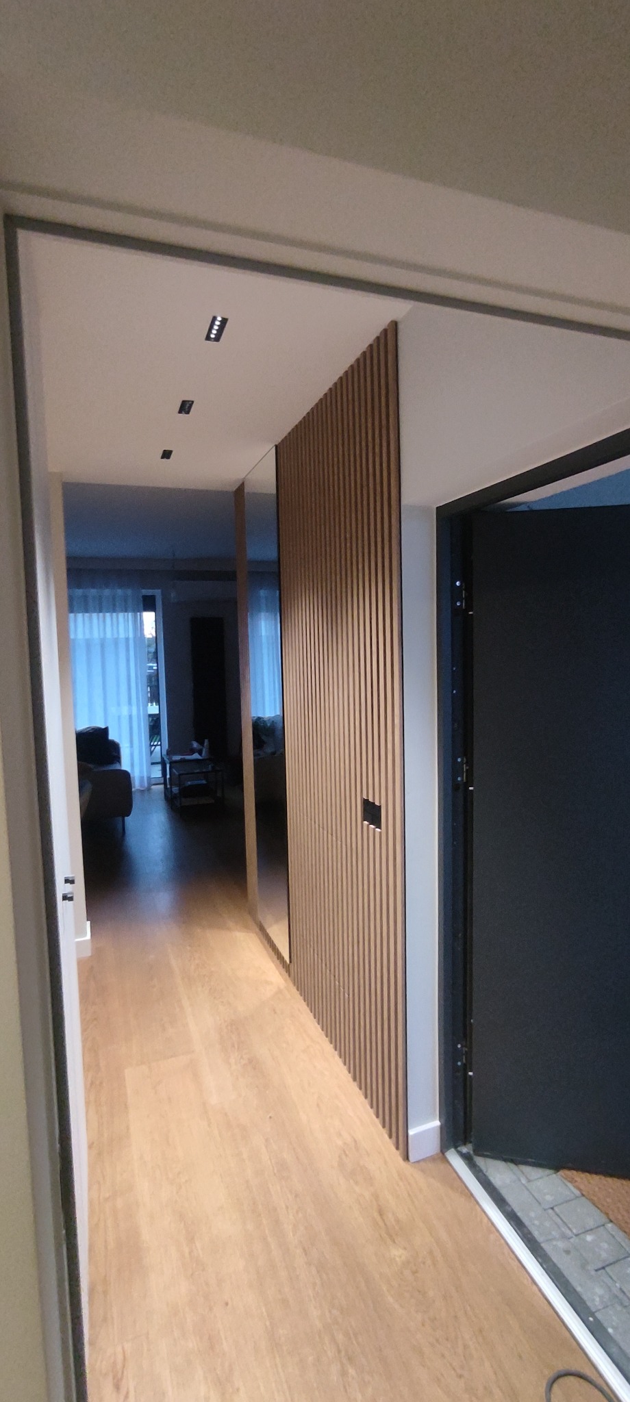 Nowoczesny przedpokój z drewnianą ścianką działową i lustrem. Jasne panele podłogowe, minimalistyczne oświetlenie sufitowe. Widok na salon przez otwarte drzwi.