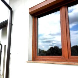 P.U.H Alex - Drewniane okno z roletą zwiniętą w kasecie, zamontowane w białej ścianie domu. W szybie odbija się pochmurne niebo i drzewa.