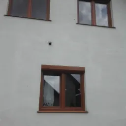 Fasada budynku z trzema oknami z brązowymi ramami i zwiniętymi roletami zewnętrznymi w kolorze brązowym. W oknach odbija się niebo i widoczne są zasłony.