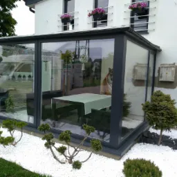 Szklana zabudowa tarasu z ciemnoszarą ramą, otoczona białymi kamieniami i niskimi iglakami, widok z zewnątrz na stół i krzesła wewnątrz, odbicie otoczenia w szybach.