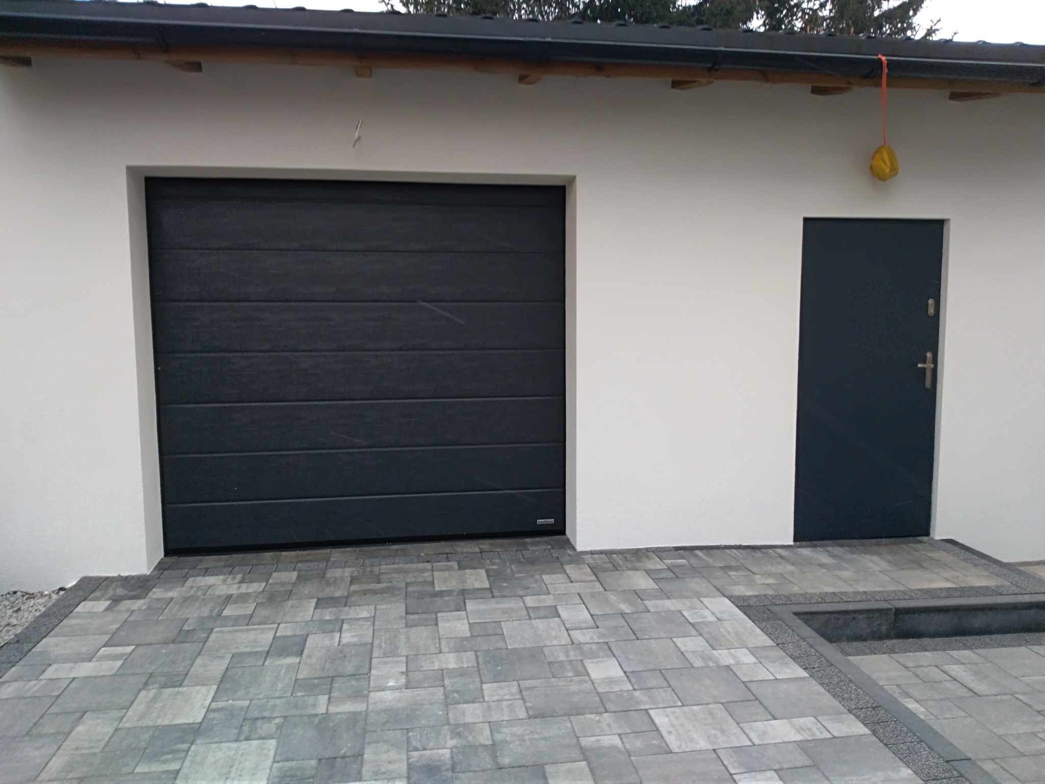 Nowoczesny garaż z szarą segmentową bramą i ciemnymi drzwiami wejściowymi, wyłożony kostką brukową w odcieniach szarości, z widocznym fragmentem dachu i żółtą osłoną na rynnie.