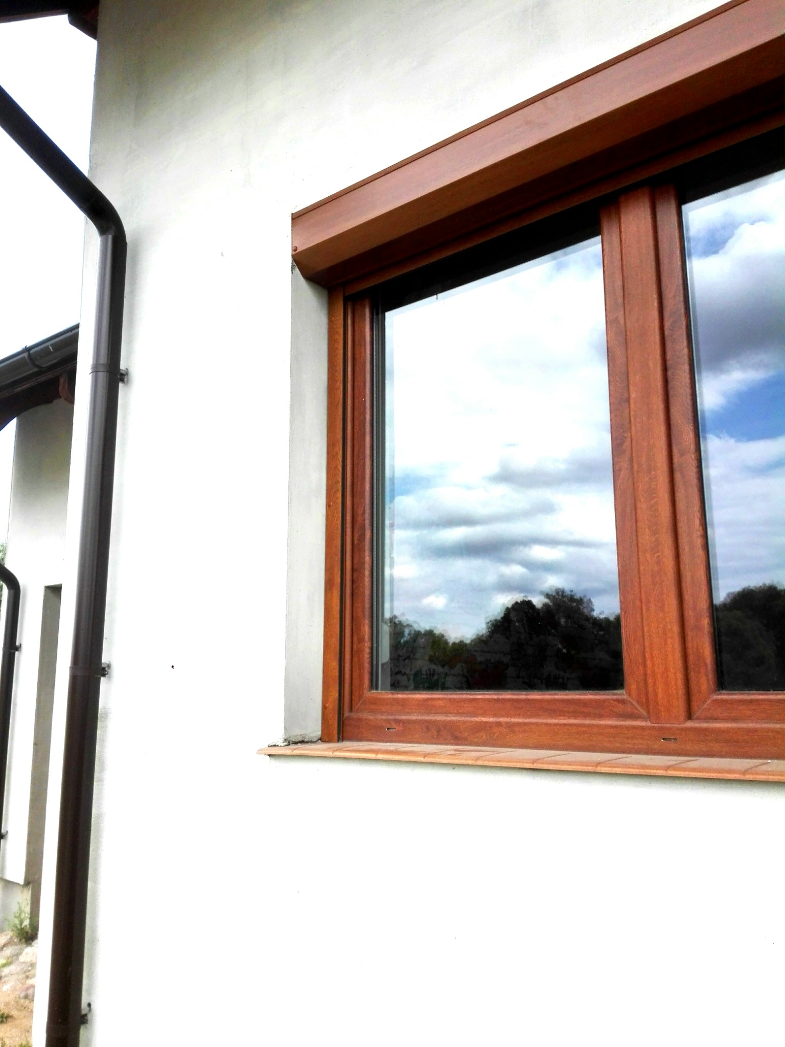 Drewniane okno z roletą zwiniętą w kasecie, zamontowane w białej ścianie domu. W szybie odbija się pochmurne niebo i drzewa.