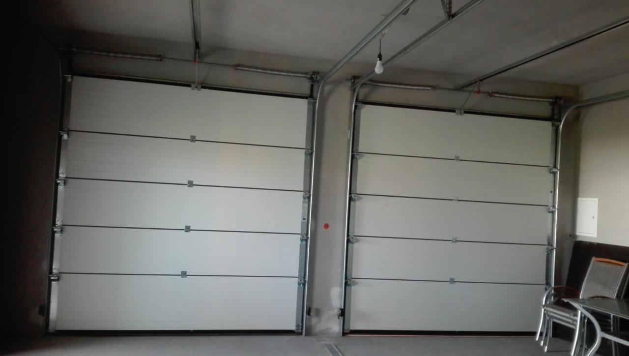 Dwie białe segmentowe bramy garażowe z widocznymi prowadnicami i sprężynami, oświetlone pojedynczą żarówką zwisającą z sufitu. W rogu widoczne krzesła i stolik.