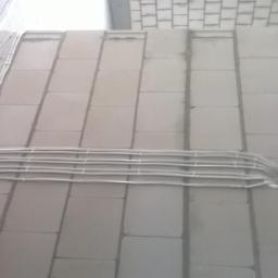 Wojciech Siwko - Białe przewody elektryczne ułożone równolegle i przymocowane do surowej ściany z bloków betonowych. Widoczne mocowania przewodów. Przewody prowadzą do otworu w ścianie.