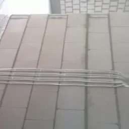 Białe przewody elektryczne ułożone równolegle i przymocowane do surowej ściany z bloków betonowych. Widoczne mocowania przewodów. Przewody prowadzą do otworu w ścianie.