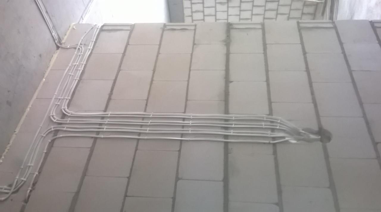 Białe przewody elektryczne ułożone równolegle i przymocowane do surowej ściany z bloków betonowych. Widoczne mocowania przewodów. Przewody prowadzą do otworu w ścianie.